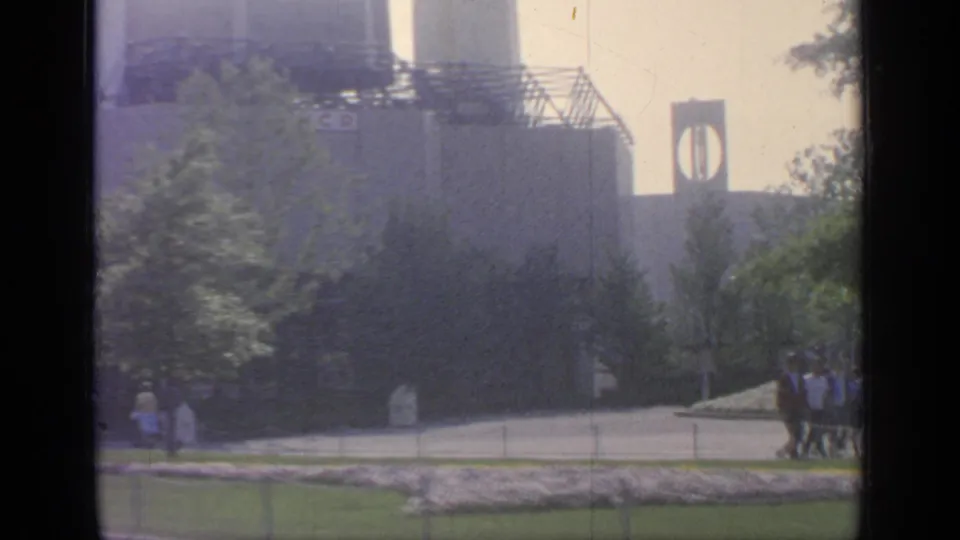 1964 Flushing Meadows fairground view with modern architecture, trees, a paved path, and visitors walking in the distance.