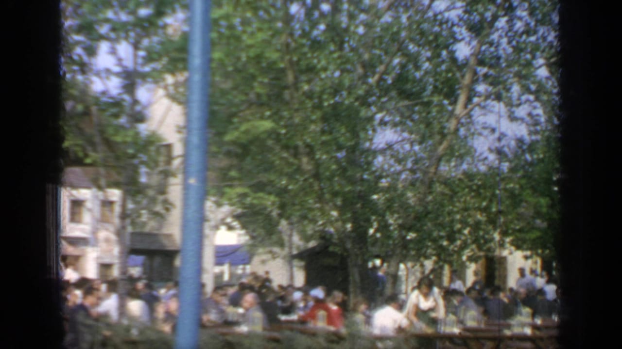 Crowds sit at outdoor picnic tables under trees, seen through dark foreground frame edges.