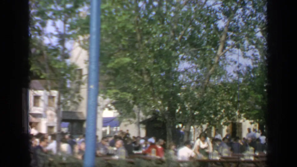 1964 Flushing Meadows outdoor dining area with people seated at long tables beneath trees, viewed through a dark foreground frame.