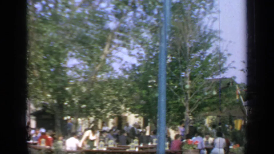 Busy 1964 Flushing Meadows dining area with people at wooden tables, blue poles, trees, and a striped canopy in the background.