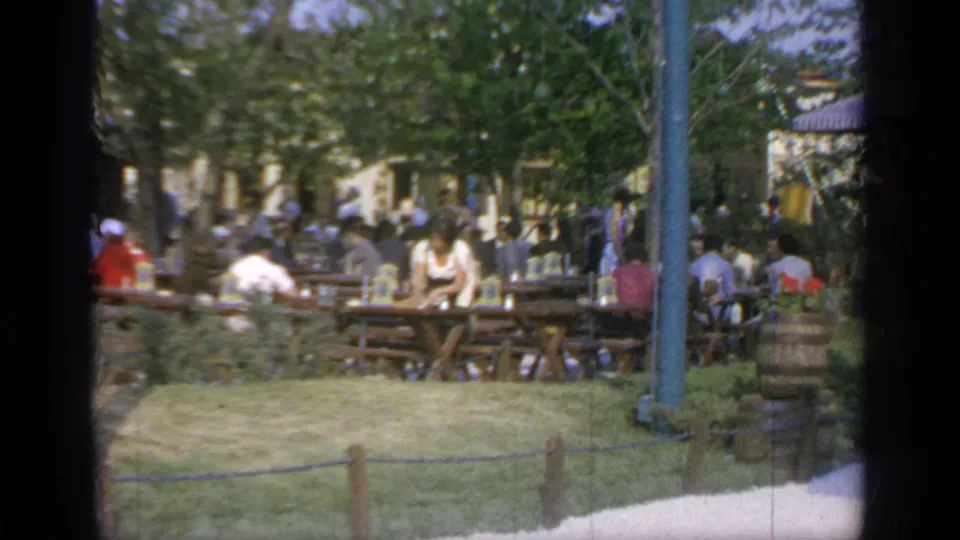 1964 Flushing Meadows food court view with diners at long tables, grass in front, trees overhead, and fairground fixtures nearby.