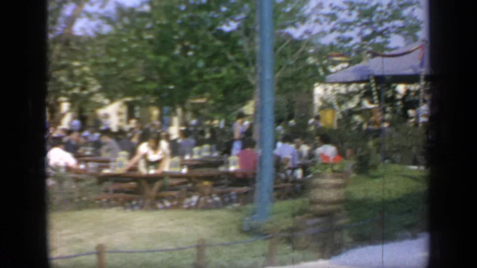 Final frame of the 1964 Flushing Meadows outdoor dining scene, showing seated visitors, wooden tables, trees, blue poles, and a striped canopy.