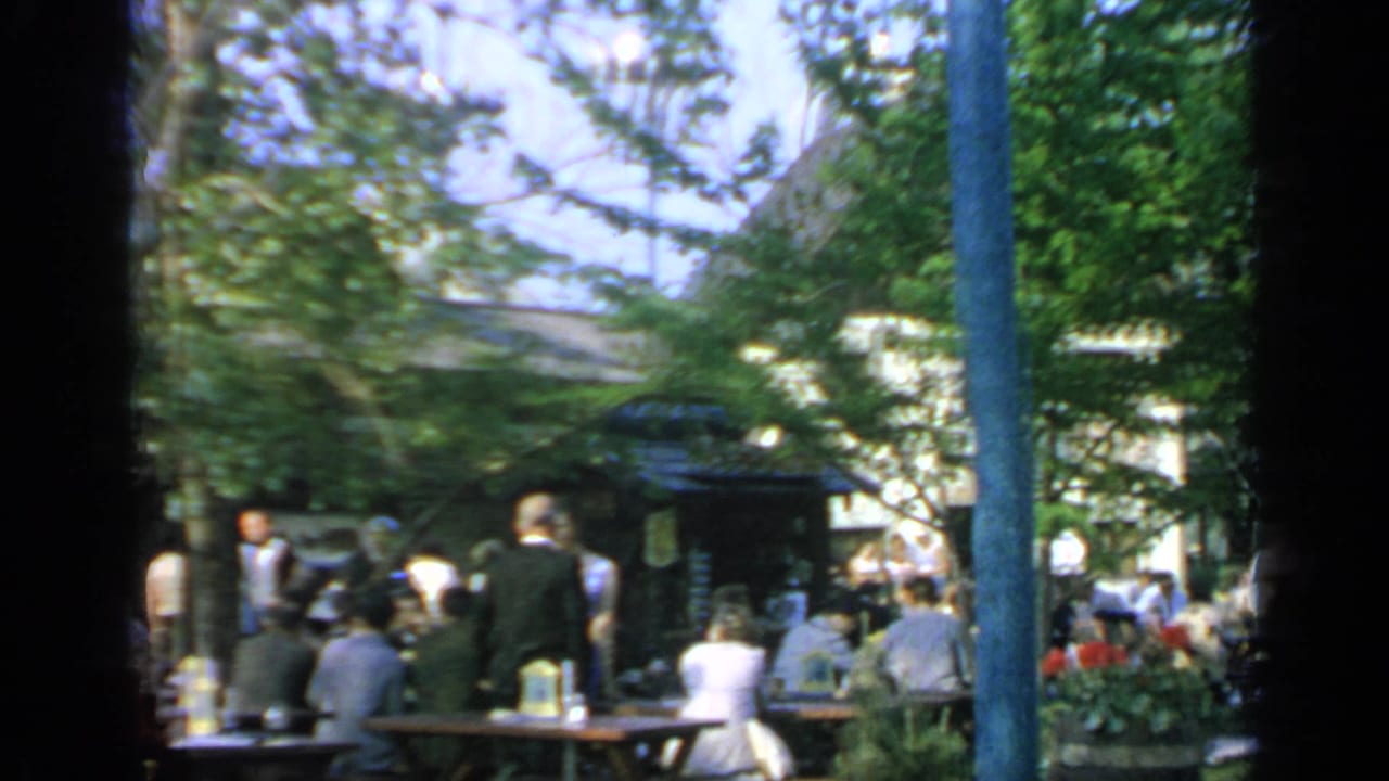 Visitors sit and stand at picnic tables under trees at an outdoor fair area.