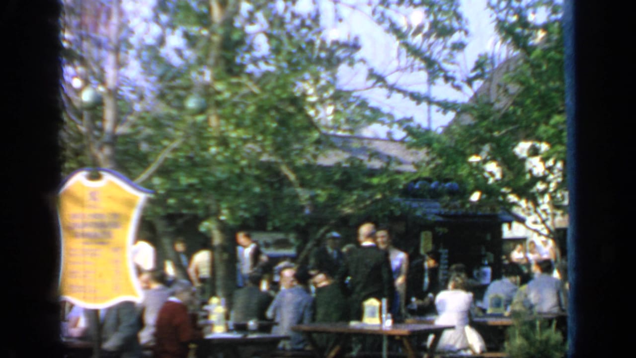 A yellow sign stands near seated and standing visitors in a shaded fair area.