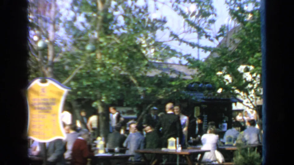 1964 Flushing Meadows frame with a yellow posted sign, picnic tables, and visitors under trees.
