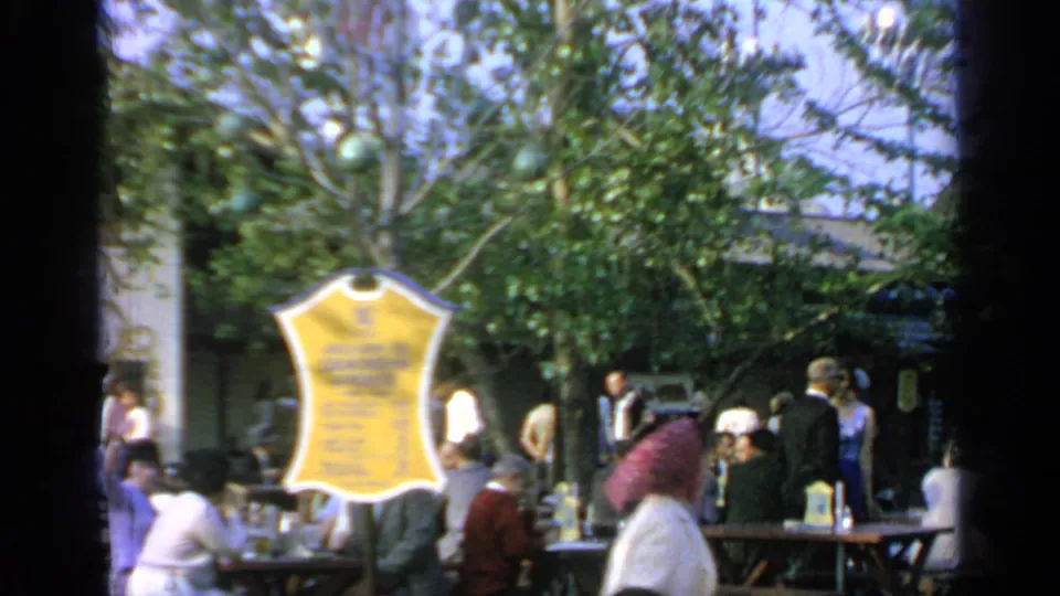 1964 Flushing Meadows home-movie frame showing a yellow sign, shaded tables, and fair visitors.
