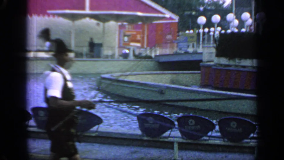 At Flushing Meadows in 1964, a man in dark shorts passes blue seating near a red World's Fair structure and plaza fountain area.