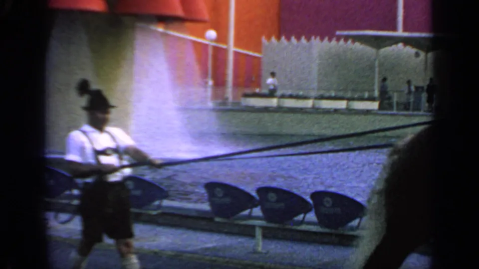 The visitor-shot frame shows a man in dark shorts near the fountain spray at the 1964 Flushing Meadows fairground plaza.