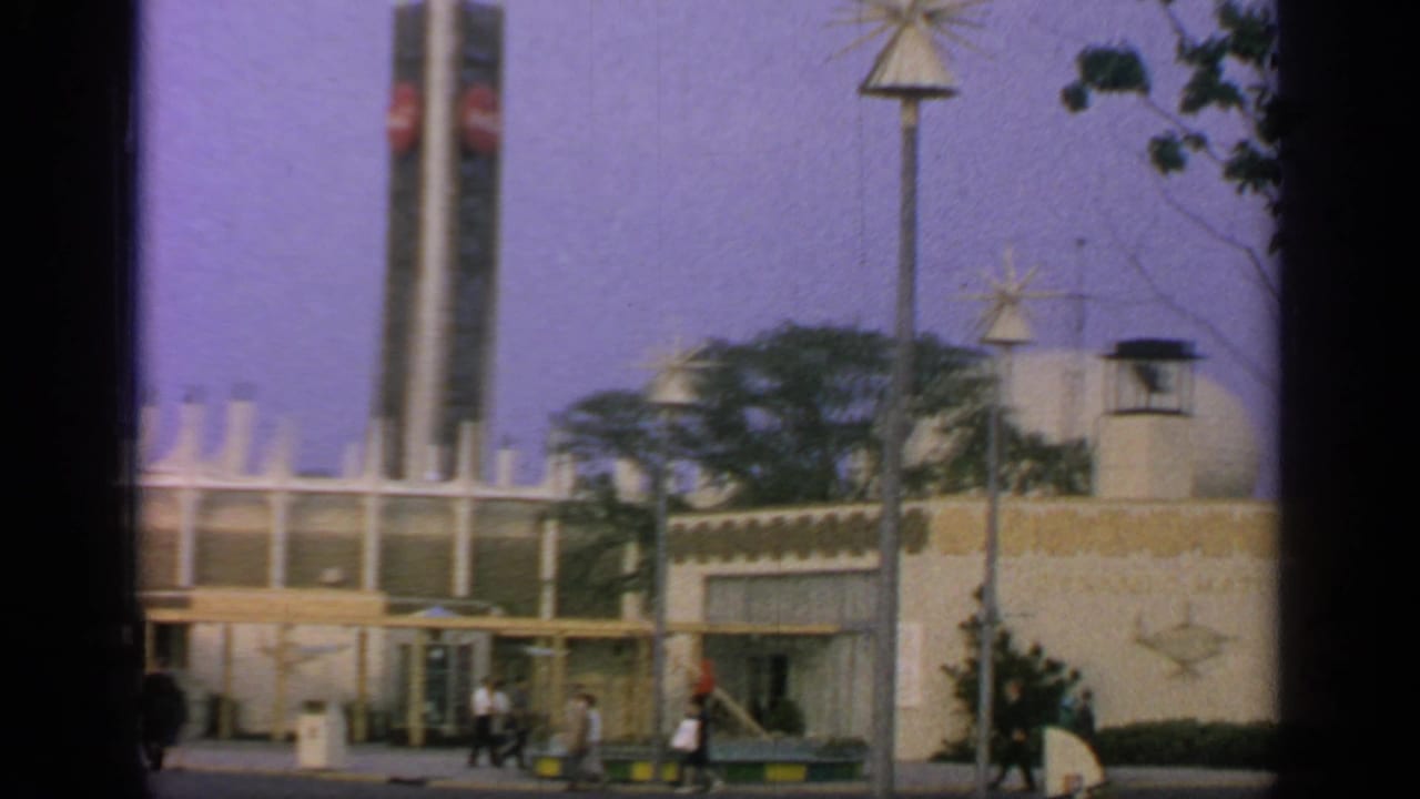 1964 Flushing Meadows fairground view with modern pavilion structures, star-shaped lamps, trees, and visitors walking past.