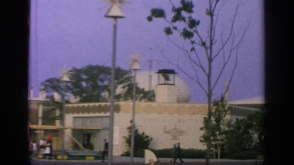 1964 Flushing Meadows exterior with a pale pavilion wall, rounded roof form, star lamps, trees, and visitors along the path.