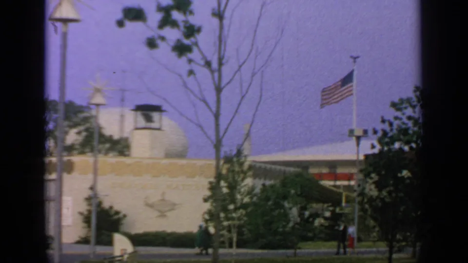 1964 Flushing Meadows view with an American flag, fairground pavilion wall, trees, star lamp, and pedestrians near landscaping.
