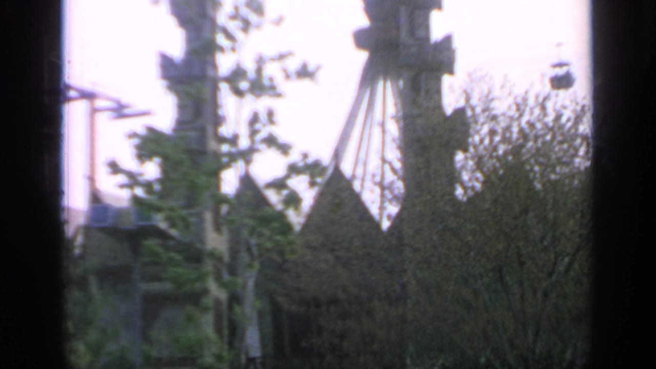 Cable car support and pavilion roof peaks rise above trees in a soft fairground view.
