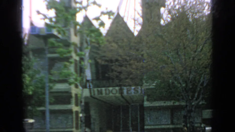 1964 Flushing Meadows frame with the Indonesia Pavilion sign, trees, poles, and tiered roof peaks partially blocked by foreground framing.