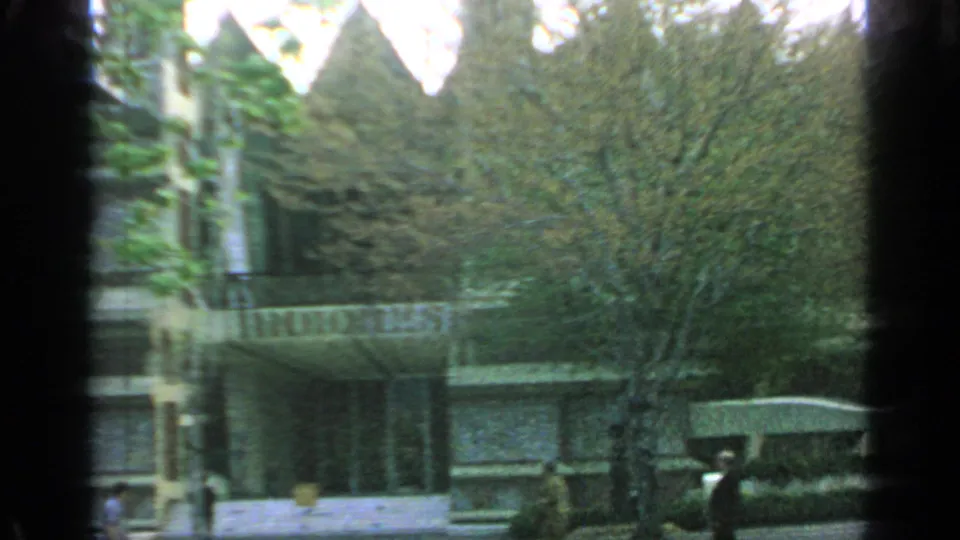 1964 Flushing Meadows fairground view with an Indonesia pavilion sign, trees, low walls, and shaded walkways seen through a handheld 8mm frame.