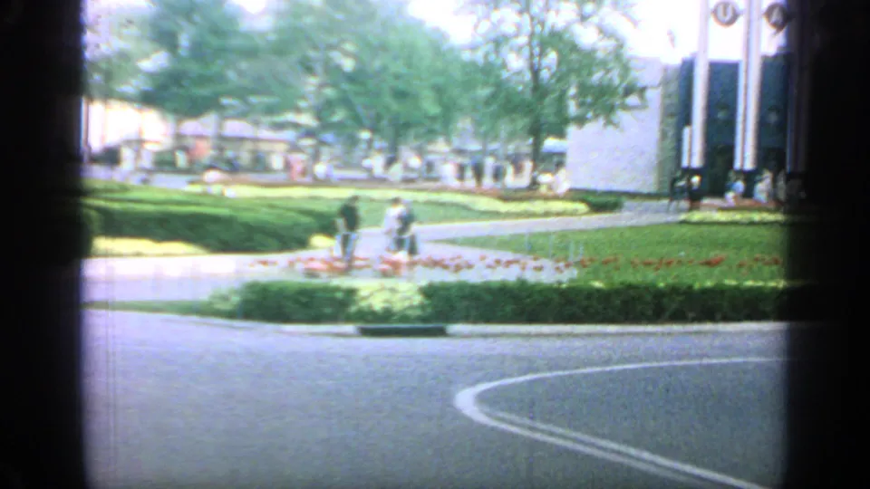 The camera looks across a curved road, lawn, plantings, and visitors toward a modern pavilion facade at the 1964 Flushing Meadows fairgrounds.