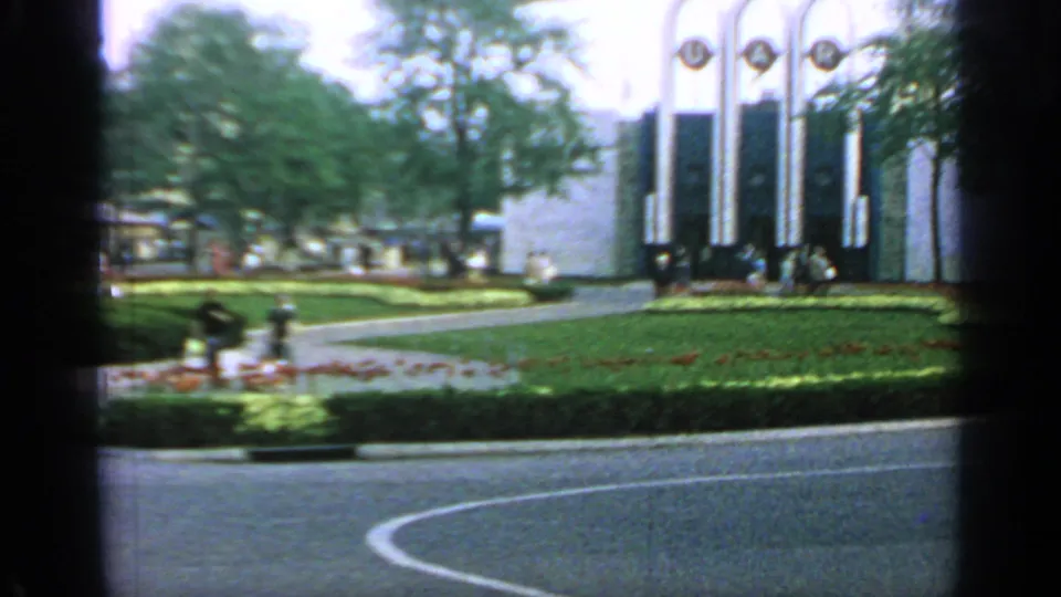 A modern pavilion marked with UAR lettering stands beyond lawns, curved paths, trees, and distant visitors in 1964 Flushing Meadows footage.