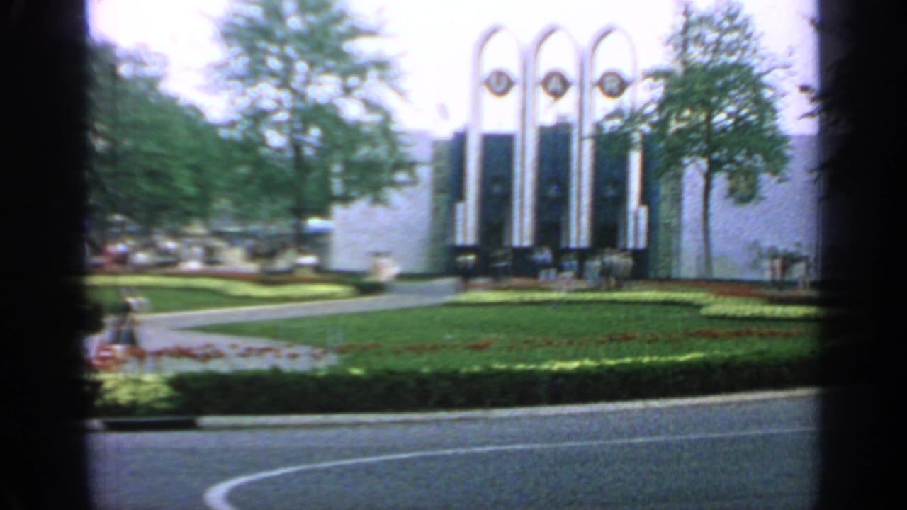 1964 Flushing Meadows fairground view with visitors, lawns, flower beds, trees, and the U.A.R. arches in soft 8mm color.