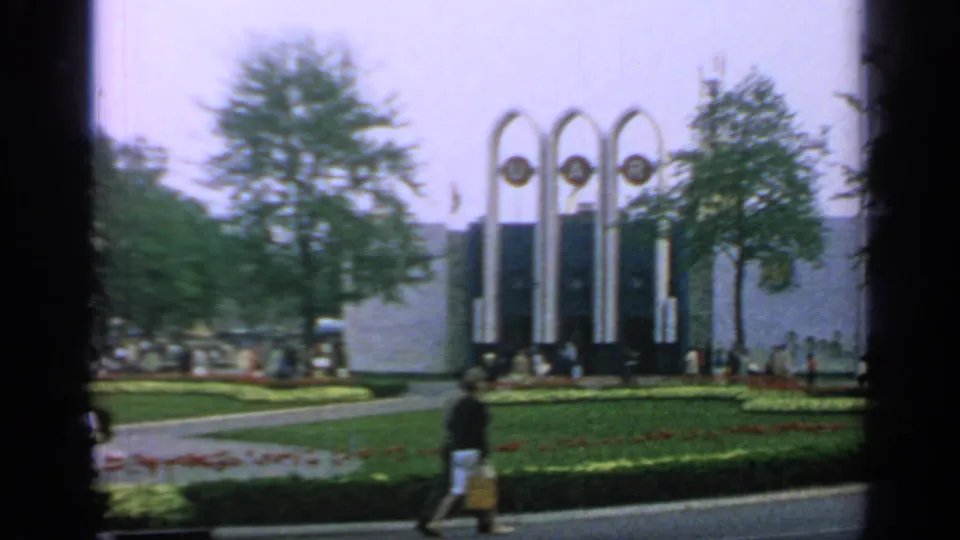 1964 Flushing Meadows scene with pedestrians, flower beds, trees, and the U.A.R. arches framed by soft home-movie film grain.
