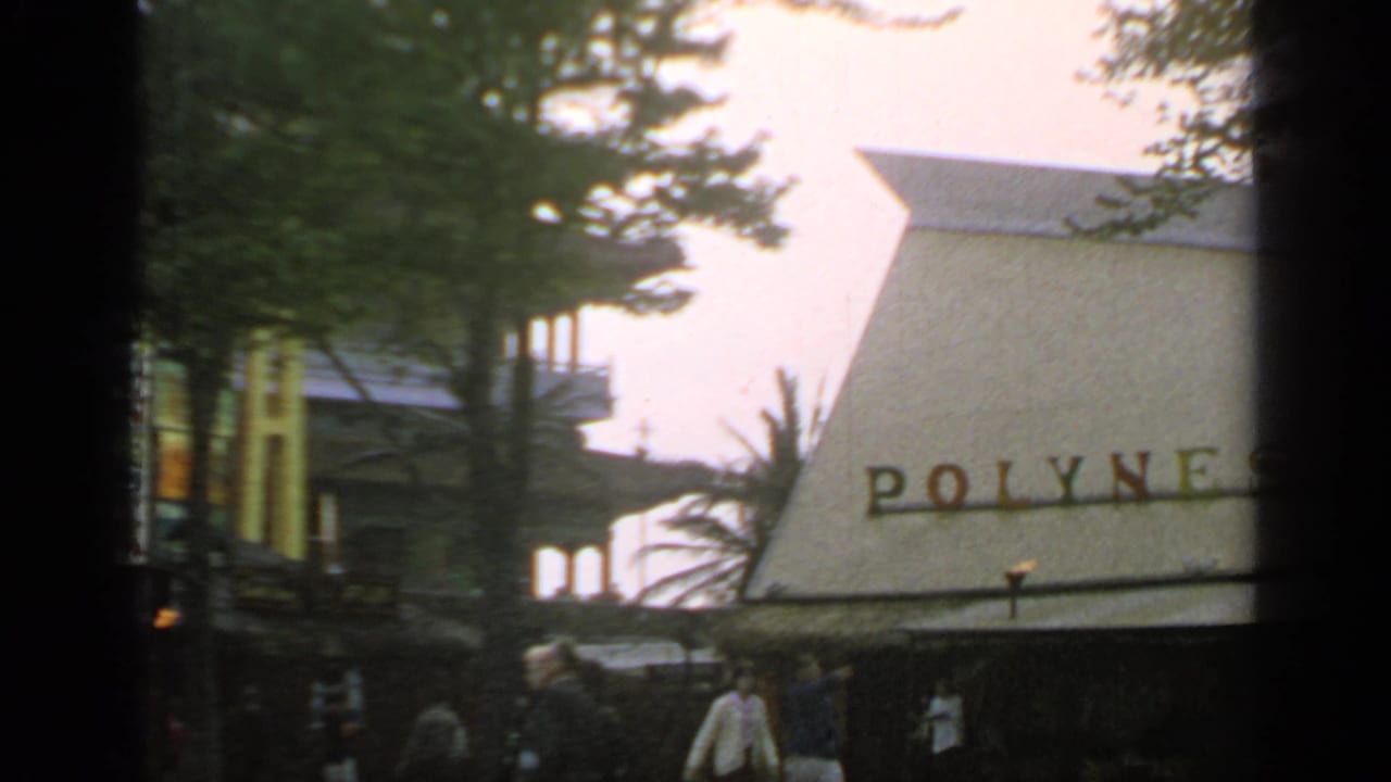 The Polynesian Restaurant sign and steep roof appear through trees as fair visitors move along the walkway below.