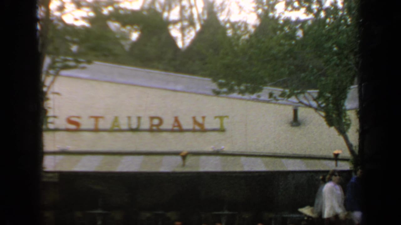 Restaurant sign on a pale building partly blocked by trees and dark foreground bands.