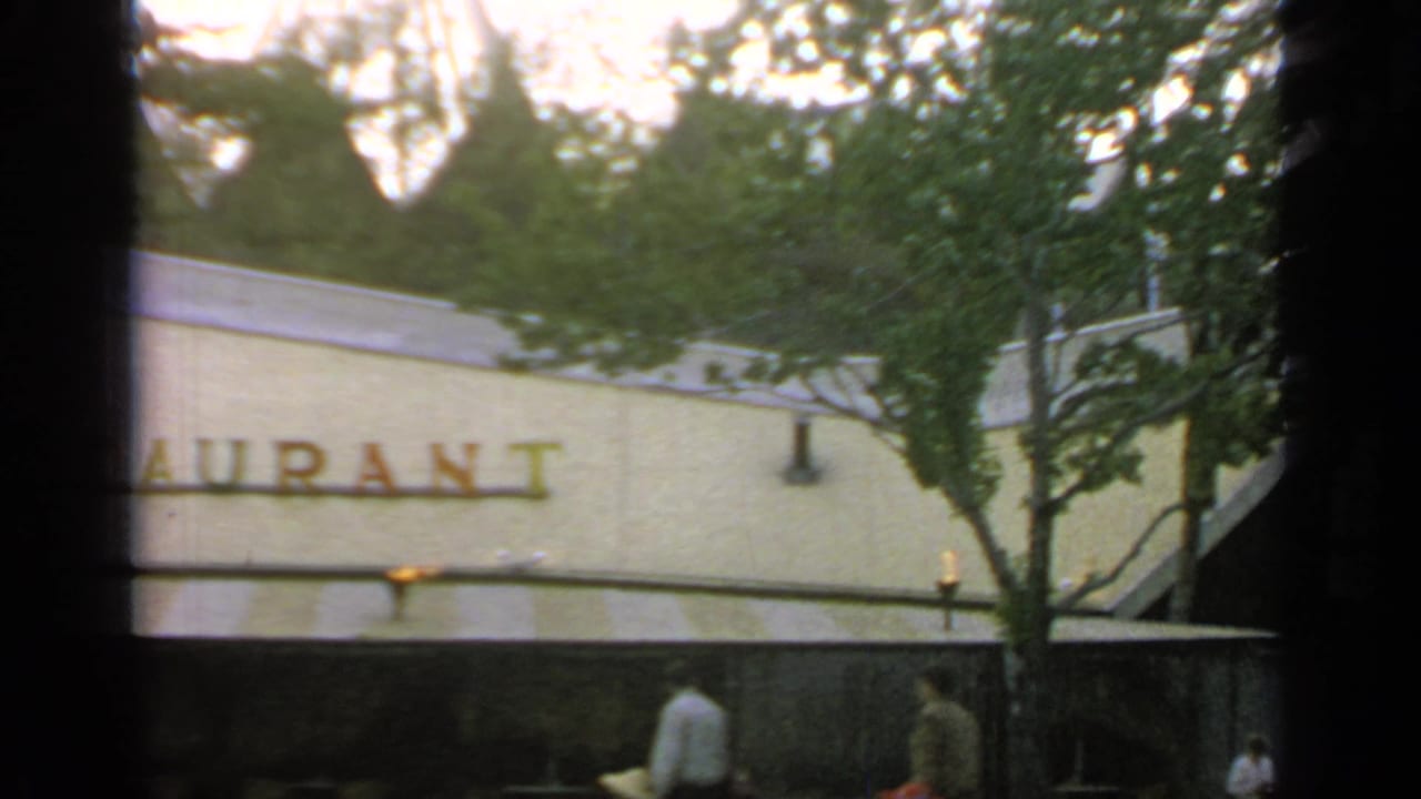 Restaurant facade with red letters, trees, awnings, and pedestrians near the lower edge.