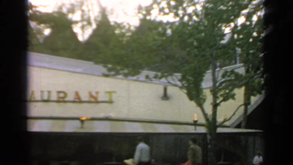 At Flushing Meadows in 1964, pedestrians pass below a partly hidden restaurant sign on a pale building.