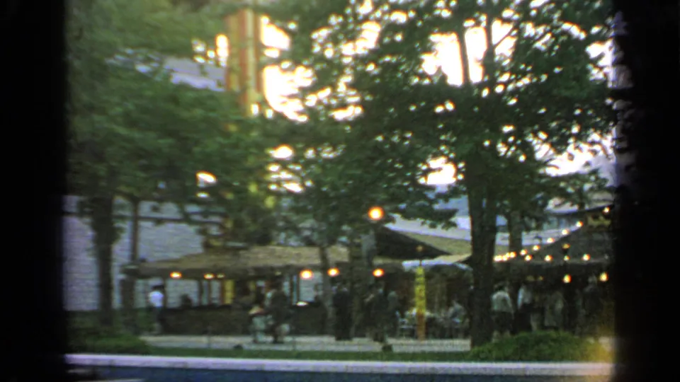 1964 fairground scene at Flushing Meadows with string lights, trees, visitors, and a shaded outdoor gathering area.