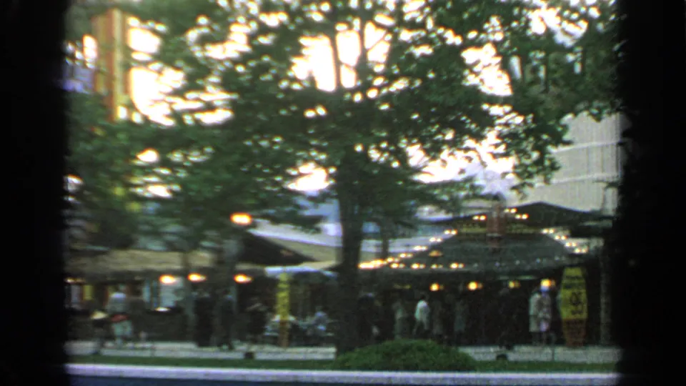 Flushing Meadows 1964 home-movie frame showing visitors near lit outdoor seating or shelter, with trees and a fair building behind.