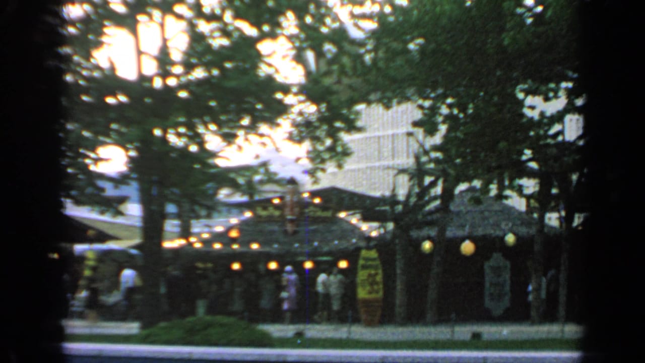 Visitors walk near a lit thatched-roof market hut at Flushing Meadows, with trees and a modern building behind it.