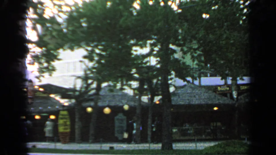 The market at Flushing Meadows appears through trees, with thatched roofing, hanging lights, signage, and a few visitors near the stalls.