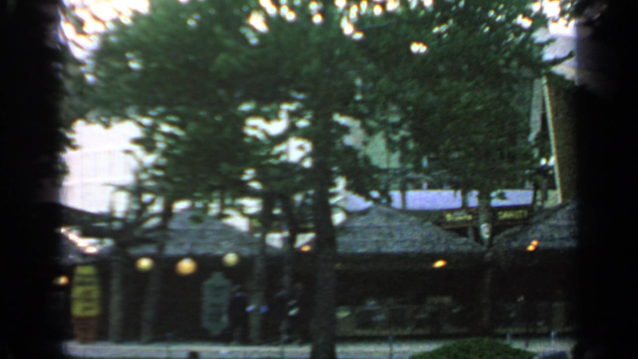 Shaded fairground walkway with trees, thatched-roof structures, lamps, and distant visitors.