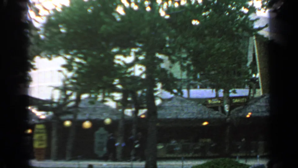 1964 Flushing Meadows fairground view with trees, low pavilion roofs, glowing lamps, and people moving through the shaded plaza.