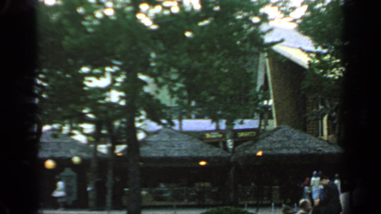 Tree-lined plaza with pavilion roofs, lamps, building edge, and small groups of visitors.