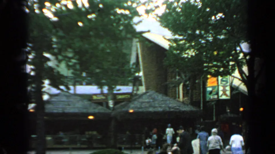1964 Flushing Meadows fairground crowd near low pavilion roofs, tree shade, lamps, and visible sign panels.