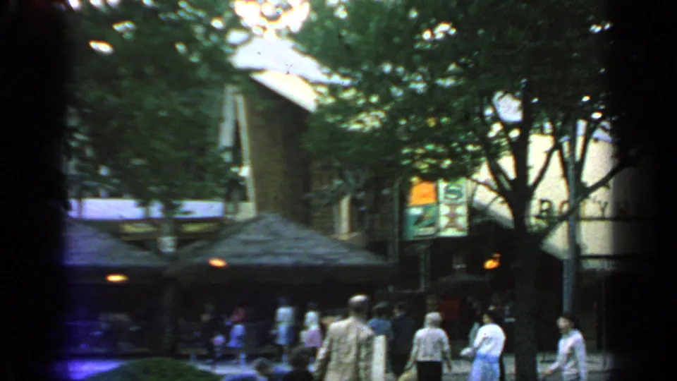 1964 Flushing Meadows fairground scene with people walking past shaded pavilion roofs, tree trunks, lamps, and visible signage.