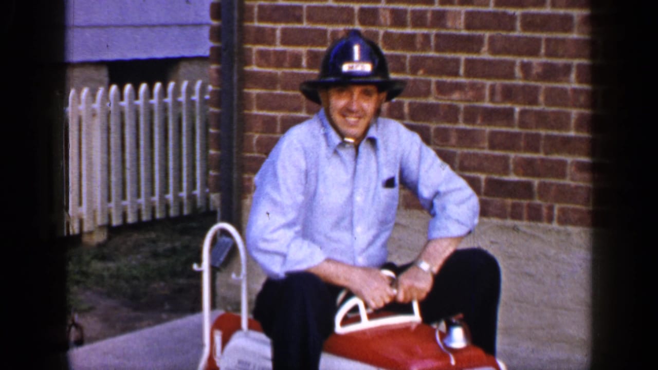 In 1965 Long Island home-movie footage, a man in a blue shirt and black firefighter-style helmet sits on a red toy fire truck.