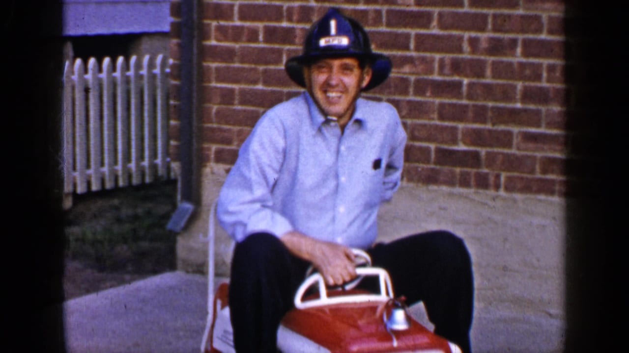 A smiling man in a blue shirt and firefighter-style helmet sits on the red toy fire truck during the 1965 home-movie scene.
