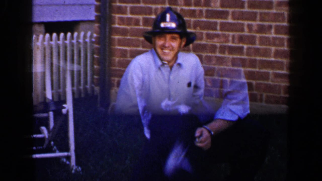 In a 1965 Long Island home-movie frame, a man in a blue shirt and firefighter helmet smiles near a red-and-white toy fire truck by brick siding.