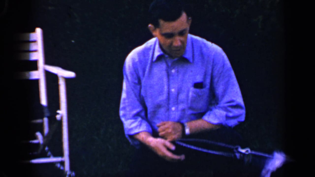 Man in blue shirt looks down while holding a leash near a small black-and-white dog.