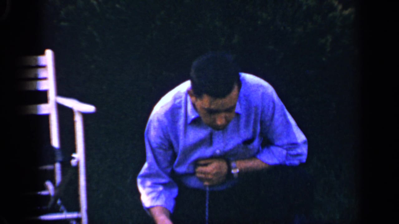 A 1965 home-movie frame shows the man bending forward on a dark lawn, with a white outdoor chair at the edge of the frame.