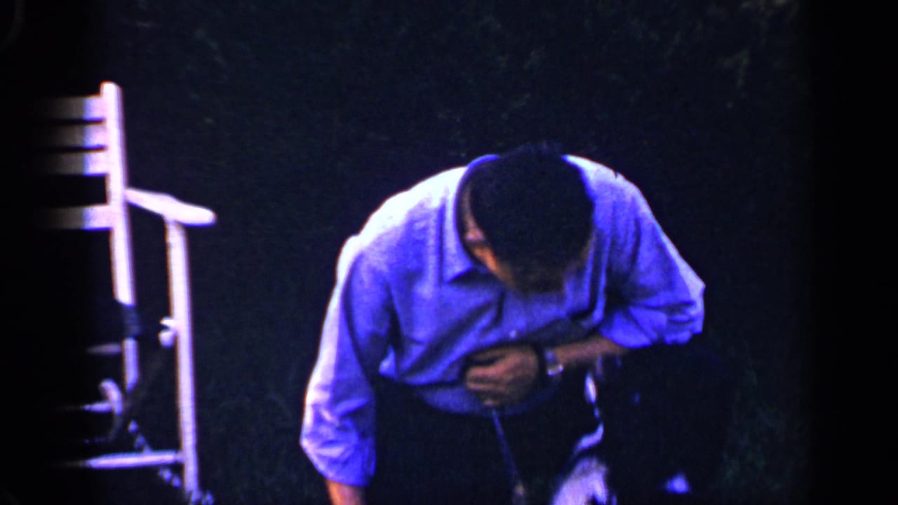 The man crouches on the lawn in the 1965 Long Island home-movie scene, reaching toward a small black-and-white dog near his knees.