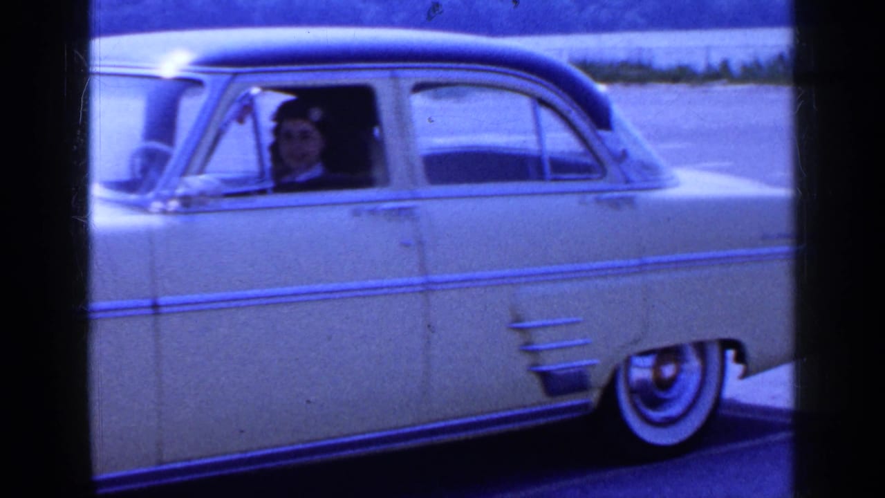 A yellow vintage car is seen in profile in a Long Island parking area, with the woman driver visible through the side window.