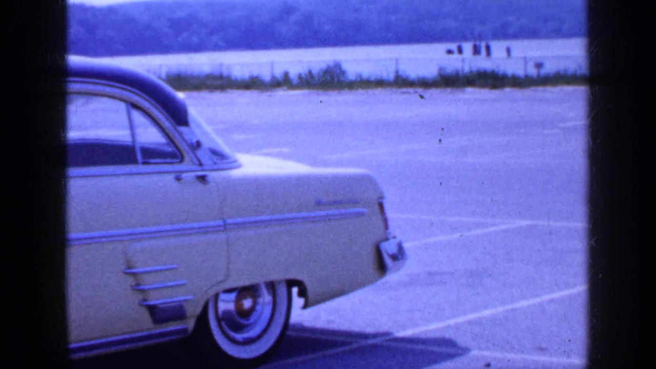 The rear half of the yellow vintage car passes out of view across painted parking spaces in the 1965 Long Island home-movie scene.