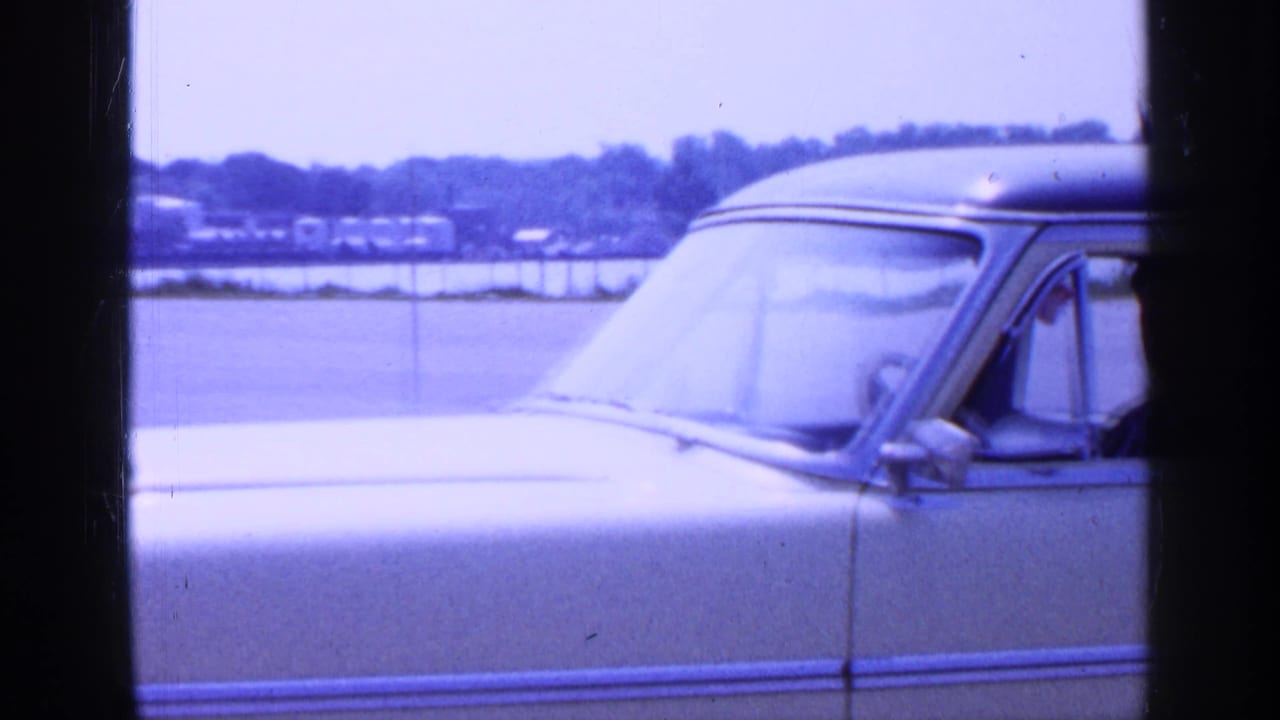 Close side view of the yellow vintage car in the Long Island parking lot, showing the windshield, side mirror, chrome trim, and driver area.