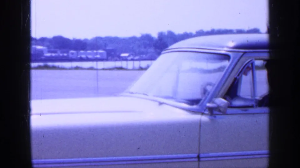 Close side view of the yellow vintage car in the Long Island parking lot, showing the windshield, side mirror, chrome trim, and driver area.