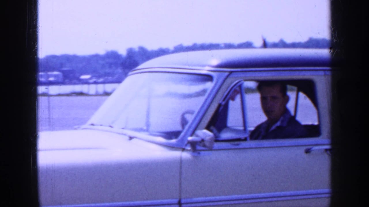 Light-colored 1960s sedan beside water with an adult male driver looking out the window.