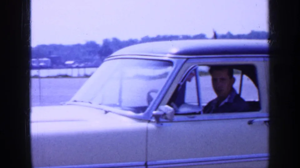 1965 Long Island home-movie frame showing a man in a light-colored sedan with dark roof near a waterfront shoreline.