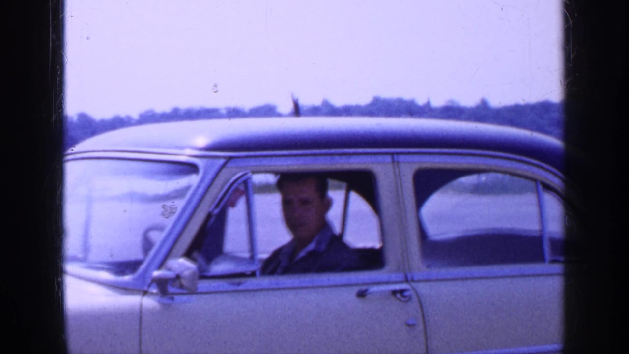 1965 Long Island frame of an adult man looking toward the camera from a parked light-colored sedan by the water.
