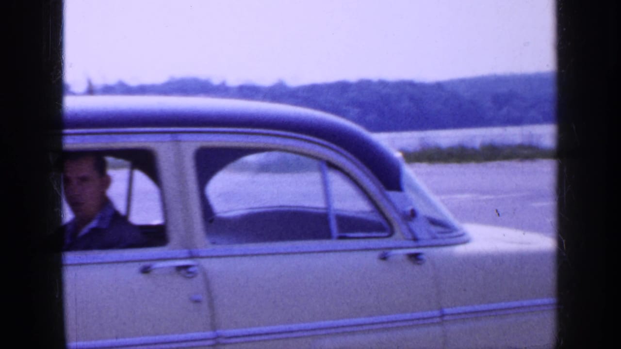 1965 Long Island home-movie frame with the sedan cropped close, showing the driver at the edge and shoreline beyond.