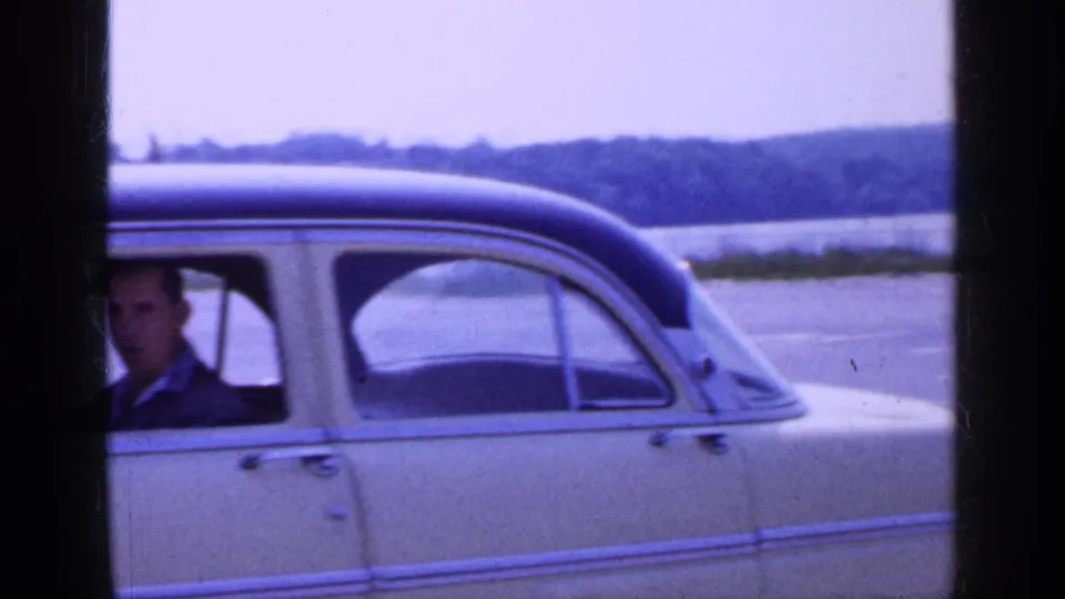 1965 Long Island home-movie frame with the sedan cropped close, showing the driver at the edge and shoreline beyond.